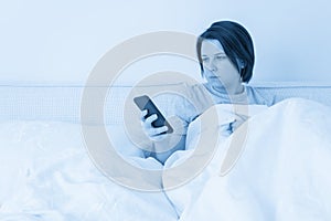 Woman using smartphone as she sits in bed covered with duvet