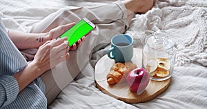 Woman using smartphone with chroma key green screen in bed during breakfast