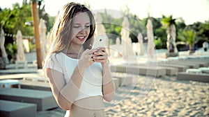 Woman using smartphone with earphones laughing on beach listening to music. Girl in bikini using