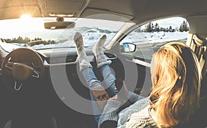 Woman using smartphone inside car with feet warm socks on dashboard - Girl relaxing in auto trip reading travel book with snow