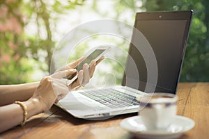 Woman using smartphone with laptop and a cup of coffee on the wooden table outdoor.