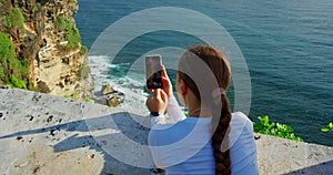 Woman using smartphone and relaxing at rocky beach. Girl taking pictures of ocean on phone. Uluwatu Bali Island
