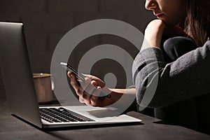 Woman using smartphone at table with laptop in dark room. Loneliness concept