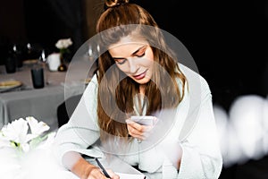 Woman using smartphone and writing in notepad in cafe with cup of coffee