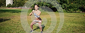 Woman using stretching band, resistance fitness rope for workout in park, doing squats, training on fresh air