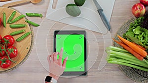 Woman using a tabletPC with a green screen at the kitchen table with fresh vegetables and fruits. Application for photo