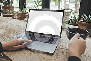 Woman using and typing on laptop with blank white desktop screen while drinking coffee on vintage wooden table