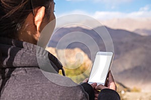 A woman using white mobile phone with blank screen while standing in front of mountain and blue sky