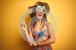 Woman on vacation wearing bikini and pineapple sunglasses over isolated yellow background smiling with happy face winking at the