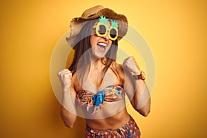 Woman on vacation wearing bikini and pineapple sunglasses over isolated yellow background very happy and excited doing winner