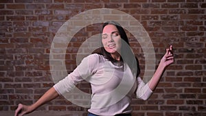 Woman is vigorously dancing with her hands high on a bricken background.