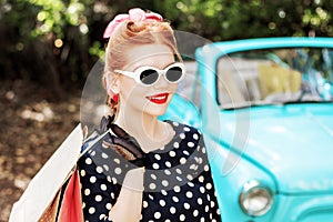 Woman in vintage clothes is holding shopping bags on background of old car.