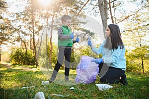 Woman volunteer and little boy picking up the plastic garbage and putting it in biodegradable trash-bag outdoors. Ecology,