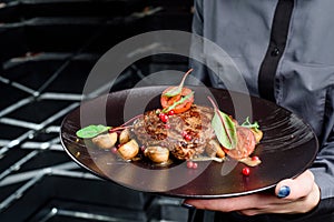 Woman waiter holding a dark round plate with grilled juicy steak with grilled mushrooms, tomatoes and cranberries.