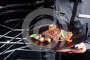 Woman waiter holding a dark round plate with grilled juicy steak with grilled mushrooms, tomatoes and cranberries.