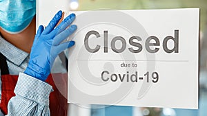 Woman waiter in protective medical mask hangs closed sign on front door of cafe. Sign closed due to Covid 19 on shop