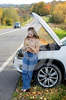 A woman waits for assistance near her car broken down on the road side. Woman with a broken car with open hood. Upset