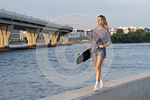 Woman walk along on river embankment hold longboard enjoy summer sunset in city. Urban lifestyle