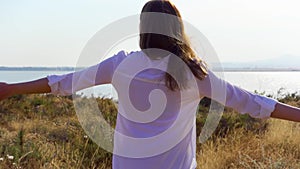 Woman walk and raise arms up in meadow near lake. Female traveler outstretching hands in slow motion