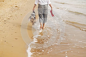Woman walking barefoot on beach, back view of legs. Young girl relaxing on sandy beach, walking with shoes and bag in hands.