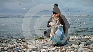 Woman walking on the beach, picking up trash, and throwing it in a garbage bag. Concept of beach cleanup, environmentalism, and