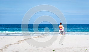 Woman walking the beach to ocean carrying towel and bags.