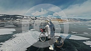 Woman is walking beside bicycle on the ice. The girl is dressed in a silvery down jacket, backpack and helmet. Ice of