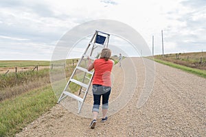 Woman walking down a rural road with a step ladder