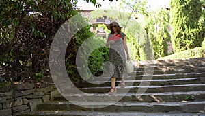 Woman walking down the stairs of a public park in a dress and straw hat on a sunny day.