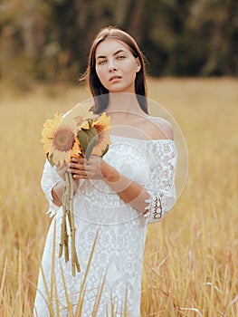 Woman walking in a field and holding a bouquet of sunflowers. Portrait of charming woman wearing white dress. Lifestyle