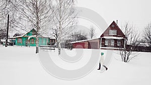 Woman walking near old wooden houses in winter snowfall weather in village.