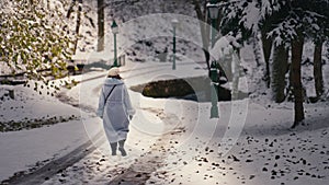 Woman Walking on a Snowy Path in the Park Next to the River, Trees, and Lamps