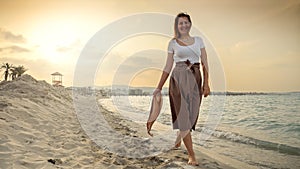 Woman walking towards the sunset on a sandy sea beach, holding a silk scarf and capturing the essence of happiness and positivity