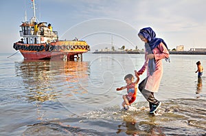 Woman walks along shore of Persian Gulf with her child.