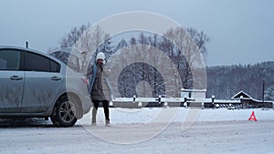 A woman walks around a broken car and calls from her smartphone. Winter, snow is falling. Breakdown on the road. auto