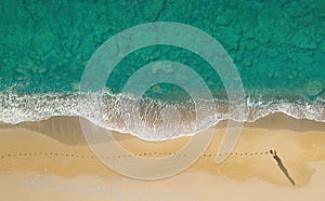 Woman walks on the beach along breaking waves leaving footprints on a sand, aerial view directly above