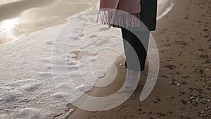 Woman walks in boots along sea foam blown by wind on sandy beach in slow motion. Female feet in waterproof footwear go