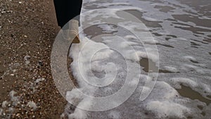 Woman walks in boots along sea foam blown by wind on sandy beach in slow motion. Female feet in waterproof footwear go