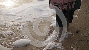 Woman walks in boots along sea foam blown by wind on sandy beach in slow motion. Female feet in waterproof footwear go