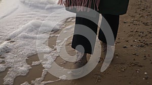 Woman walks in boots along sea foam blown by wind on sandy beach in slow motion. Female feet in waterproof footwear go