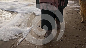 Woman walks in boots along sea foam blown by wind on sandy beach in slow motion. Female feet in waterproof footwear go
