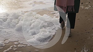 Woman walks in boots along sea foam blown by wind on sandy beach in slow motion. Female feet in waterproof footwear go