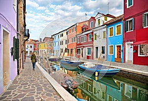 Woman walks in Burano island picturesque street with small colored houses in row, water canal with fishermans boats, c