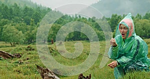 Woman walks on meadow in raincoat looks at brown horse. Landscape cloudy scenic view. Picturesque mist forest in light