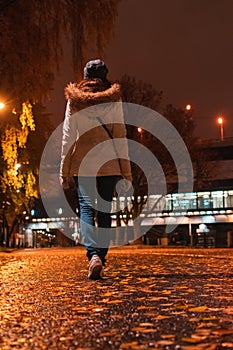 Woman walks at night in an autumn park. Rear view of young female in white coat walking away on wet alley