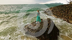 Woman walks on rock of sea reef stone, stormy cloudy ocean. Blue swimsuit dress tunic. Concept rest, tropical resort