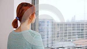 Woman walks to the kitchen and opens the blinds to look through them