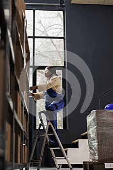Woman warehouse worker standing on ladder and taking box