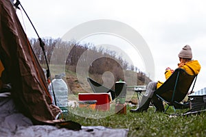 Woman in warm jacket and hat sitting on folding chairs, tourist tent and ice pack, thermal food box and water bottle. Breakfast in