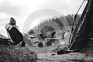 Woman in warm jacket and hat sitting on folding chairs, tourist tent and ice pack, thermal food box and water bottle. Breakfast in